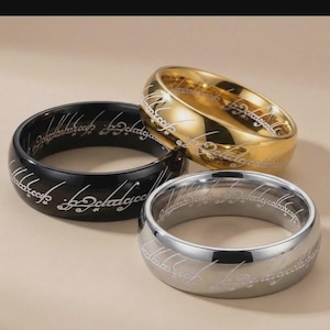 Op de afbeelding: Drie ringen in goud, zilver en zwart, elk met gegraveerde tekst. De ringen worden gepresenteerd op een neutrale achtergrond. De gouden ring heeft een glanzende afwerking, terwijl de zilveren en zwarte ringen mat zijn. Sieraden.