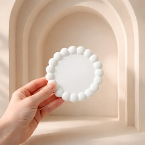 May include: A white, round coaster with a scalloped edge. The coaster is held in a hand against a neutral background. The coaster's design features a series of small, rounded bumps around the perimeter, creating a decorative border.