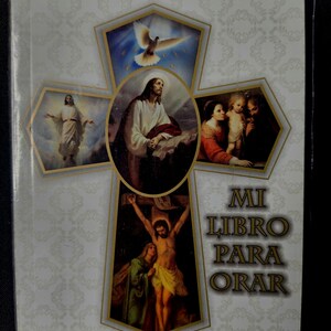 May include: A book with a cross-shaped design featuring religious imagery. The cover includes illustrations of Jesus, a dove, and other biblical scenes. The text "MI LIBRO PARA ORAR" is displayed on the cover.