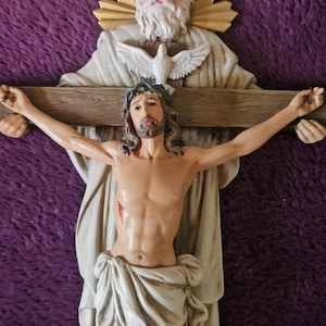 May include: A religious figurine depicting Jesus on a cross, with a figure above and a dove. The sculpture is in shades of beige and brown, with a purple background. The figure of Jesus has a crown of thorns.