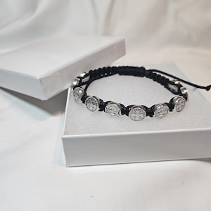 May include: A black cord bracelet featuring silver-colored, round religious medallions. The bracelet is displayed inside a white box, with the box lid slightly open. The background is a soft, white fabric.