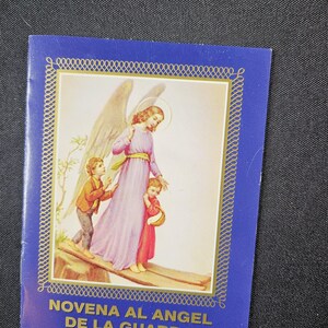 May include: A blue booklet with a gold border and the text "NOVENA AL ANGEL DE LA GUARDA." The cover features an illustration of an angel in a pink dress with two children. The angel has large wings and a halo.