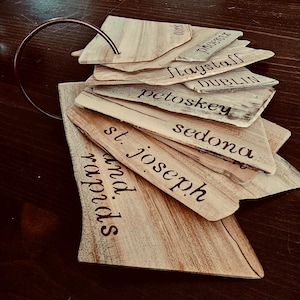 Puede incluir: Un conjunto de etiquetas de madera con nombres de ciudades grabados a fuego, unidas por un anillo de metal color cobre. Los nombres incluyen Phoenix, Flagstaff, Dublin, Petoskey, Sedona, St. Joseph y Grand Rapids.