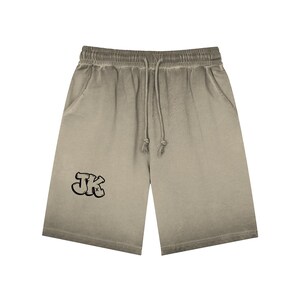 May include: Khaki-coloured shorts with a faded, ombre effect. The shorts feature a drawstring waistband and two side pockets. The letters "JK" are printed on the left leg in a black, graffiti-style font. These shorts are a casual wear item.
