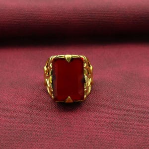 Könnte beinhalten: Ein goldfarbener Ring mit einem großen, rechteckigen, tiefroten Edelstein. Der Ring hat ein verziertes, offenes Design um den Edelstein herum, mit dekorativen Elementen an den Seiten. Der Ring ist vor einem roten Hintergrund platziert.