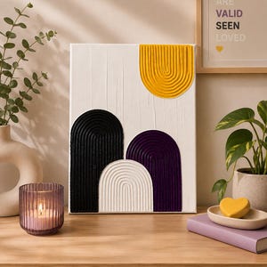 May include: Abstract textured artwork featuring arched shapes in black, white, purple, and yellow against a white backdrop. A candle in a glass holder and a framed print with the words "YOU ARE VALID SEEN LOVED" are also visible.