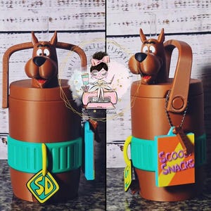 Puede incluir: Un vaso cilíndrico marrón con una cabeza esculpida de Scooby-Doo en la tapa, una pajita transparente y una banda de silicona verde azulado. Incluye una etiqueta amarilla en forma de diamante con las letras SD y una etiqueta cuadrada morada.