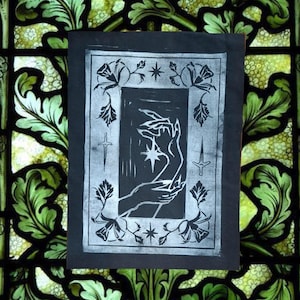 May include: A black and white linocut print on paper featuring a central rectangular frame. Inside, two hands reach towards a four-pointed star. The border is decorated with floral motifs, two daggers, and two additional stars at the top and bottom.