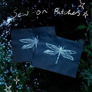 May include: Two dark blue sew-on patches with a white dragonfly design. The patches are square and feature a detailed illustration of a dragonfly. The text "Sew-on Patches" is written in white script above the patches.