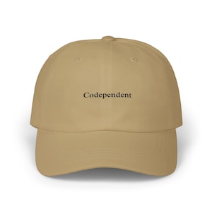 May include: A tan baseball cap with the word "Codependent" embroidered in black across the front. The cap has a curved brim and a classic six-panel design. The hat is made of a soft, durable material.