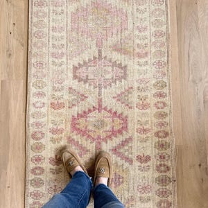 Puede incluir: Una alfombra rectangular de estilo vintage con un motivo floral central en tonos rosa y beige. La alfombra tiene un aspecto desgastado, con colores descoloridos y una textura desgastada. La alfombra está colocada sobre un suelo de madera clara. Una persona lleva vaqueros azules y mocasines marrones.