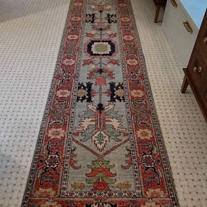 May include: A long, rectangular runner rug featuring a light blue central field with a symmetrical pattern of geometric and floral motifs in shades of red, navy blue, green, and cream, framed by a wide, intricate border in red and dark blue tones.