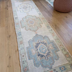 Op de afbeelding: Een lang, smal loper-tapijt met een crèmekleurige basis en een herhalend patroon van geometrische medaillons in tinten lichtblauw, saliegroen en gedempt oranje. Het tapijt heeft een decoratieve rand met kleine, kleurrijke vierkante motieven.