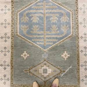 Op de afbeelding: Een rechthoekig vloerkleed met een saliegroene achtergrond en een grote lichtblauwe zeshoek in het midden met een gestileerd beige boommotief. De rand en het veld zijn versierd met kleine beige en blauwe geometrische kruis- en ruitpatronen.