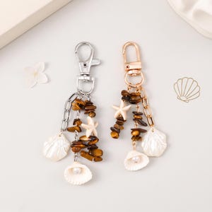 Op de afbeelding: Twee sleutelhangers met zilver- en goudkleurige karabijnsluitingen. Elke sleutelhanger bevat een ketting, bruine tijgeroogsteenfragmenten, een kleine witte zeesterbedel en een witte schelpbedel met een parelmoerkleurige kraal.
