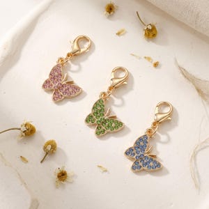 May include: Three gold-tone metal butterfly charms, each featuring a lobster claw clasp. The butterflies are encrusted with small, sparkling rhinestones in pink, green, and blue, respectively, creating a vibrant and detailed accessory set.