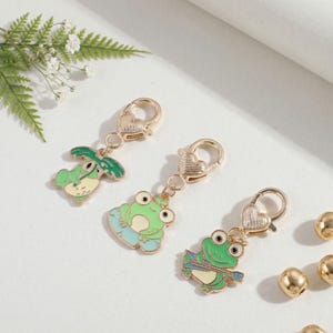 May include: Three gold-toned metal charms featuring green frog designs, each attached to a heart-shaped lobster clasp. One frog holds a leaf, one sits on a lily pad, and one holds a blue oar. The charms are finished with enamel detailing.