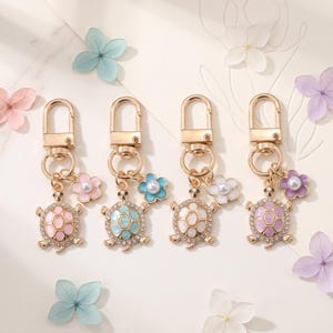 May include: Four gold-toned metal keychains featuring turtle charms with enamel shells in pastel pink, light blue, white, and lavender. Each turtle is accented with clear rhinestones and a small flower charm with a central white faux pearl bead.