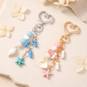 May include: Two heart-shaped lobster clasp keychains. One is silver-toned with blue beads, a white shell, and a blue starfish charm. The other is gold-toned with pink beads, a white shell, and a pink starfish charm, both featuring metal chains.