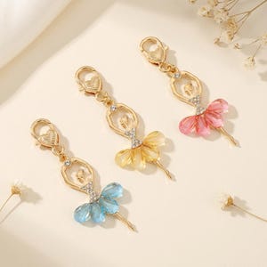 May include: Three gold-toned metal ballerina charms with lobster claw clasps shaped like hearts. Each figure features a clear rhinestone bodice and a skirt made of translucent teardrop-shaped beads in light blue, yellow, and pink colors.