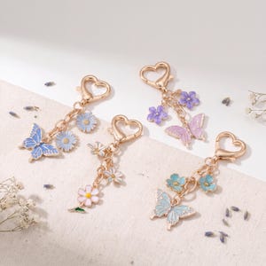 Puede incluir: Cuatro llaveros decorativos con cierres en forma de corazón. Cada llavero presenta un dije de mariposa y flor en tonos de azul, morado y rosa. Las mariposas tienen patrones de alas detallados y las flores tienen pétalos delicados. Los llaveros son dorados.