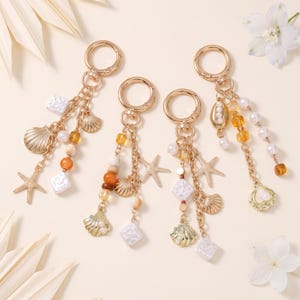 May include: Four gold-toned metal keychains featuring a circular clasp and dangling charms. The charms include gold-colored starfish, seashells, white square beads, and translucent amber and white spherical beads arranged on delicate gold chains.