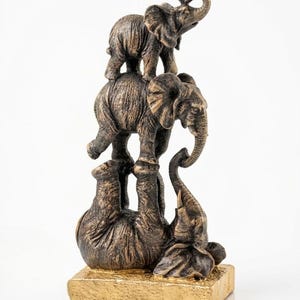 May include: A bronze-coloured elephant figurine featuring three elephants arranged in a stack. The base is a gold-coloured rectangle. The elephants are in different poses, with one standing on its head.