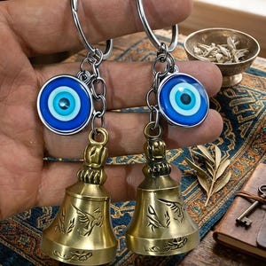 以下が含まれることがあります： シルバーのリング、チェーン、真鍮のベルが付いた2つのキーホルダー。各キーホルダーには、青と白の邪視チャームが付いています。ベルには彫刻が施されています。キーホルダーは手で持たれています。