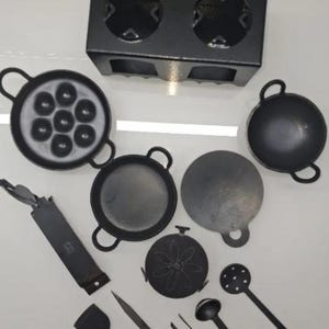 Könnte beinhalten: Eine Sammlung schwarzer Kochutensilien und -werkzeuge aus Gusseisen, darunter Pfannen, ein Kochfeld, Spatel und Schöpflöffel. Die Pfannen variieren in Form und Größe, einige weisen mehrere kreisförmige Formen auf. Die Utensilien haben lange Griffe.