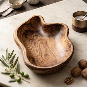 Op de afbeelding: Een houten kom met een organische vorm, met een rijke, natuurlijke houtnerf. De kom is omringd door kleine keramische kommen, een zoutvat van messing en hout, olijftakken met groene olijven en walnoten. De items zijn gerangschikt op een marmeren oppervlak.