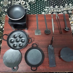 以下が含まれることがあります： 小さな黒い金属製の調理器具と調理器具のコレクション。小さなストーブ、フライパン、おたま、ヘラ、トングなど、木製の表面に配置されています。このセットは、子供のおもちゃのキッチン用であるようです。