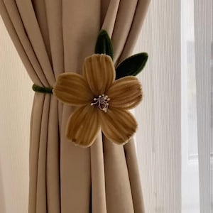 May include: A decorative curtain tieback featuring a large, golden-yellow flower with a central cluster of white beads. The flower is attached to a green, velvet-covered cord, holding beige curtains. Green leaves accent the flower.