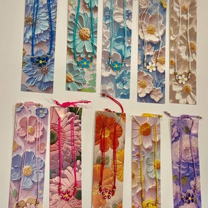 May include: A collection of floral-themed bookmarks with matching necklaces. Each bookmark features a different color scheme, with embossed flower designs. The necklaces have a flower pendant that complements the bookmark's color.