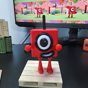 May include: A red square-shaped character figure with a single black antenna, a large circular eye with a black pupil, and a wide black mouth with white teeth. The figure has thin red arms and legs and stands on a light-colored wooden pallet.