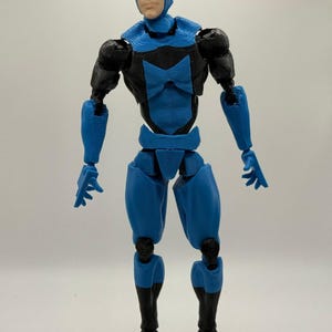 Puede incluir: Figura de acción azul y negra de un superhéroe con máscara. La figura tiene un traje azul y negro con articulaciones. La figura está de pie sobre un fondo liso.