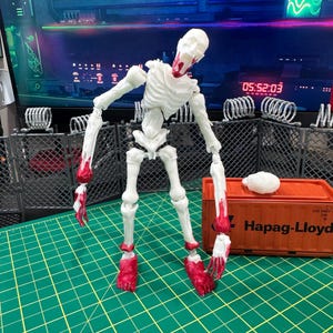 May include: A white articulated skeleton action figure with red painted details on the jaw, hands, and feet. The figure stands next to an orange Hapag-Lloyd shipping container model and a separate white skull piece on a green cutting mat.
