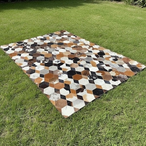 Może przedstawiać: Prostokątny dywan z geometrycznym wzorem brązowych, czarnych, białych i szarych sześciokątnych kształtów. Dywan wykonany jest ze skóry krowiej i jest prezentowany na zielonym trawniku.