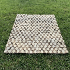 Op de afbeelding: Een rechthoekig tapijt met een geometrisch patroon van onderling verbonden zeshoeken in tinten wit, beige en bruin. Het tapijt ligt op groen gras en toont zijn ontwerp en textuur.