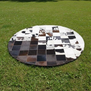 May include: A round cowhide rug with a patchwork design. The rug features a checkerboard pattern of black, white, brown, and gray squares. The rug is placed on a green grassy surface.