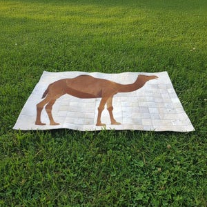 Puede incluir: Una alfombra rectangular con un diseño de camello. El camello es marrón y beige, sobre un fondo blanco con un patrón de cuadrícula. La alfombra está colocada sobre hierba verde, creando un contraste de colores y texturas.