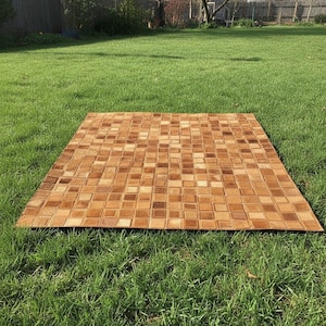 Peut inclure: Un tapis rectangulaire avec un motif en mosaïque de petits carreaux carrés dans des tons de marron et de beige. Le tapis est posé sur une pelouse verte, créant un contraste de textures et de couleurs. Le tapis semble être en cuir ou en matériau similaire.