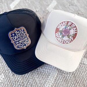 May include: Two trucker hats. One is navy blue with a patch that reads "The Darker the Night the Brighter the Stars." The other is beige with a circular patch that says "What if it all works out?" with a floral design.