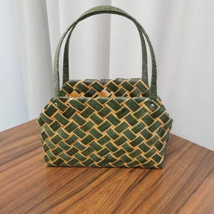 May include: A woven basket bag with a green and tan checkered pattern. The bag has two arched handles and a rectangular shape. The bag is sitting on a wooden surface.