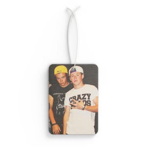 May include: Rectangular air freshener featuring a faded image of two young men. One wears a white t-shirt with "CRAZY" printed on it. The other wears a black t-shirt. Both wear baseball caps. The air freshener hangs from a white string.