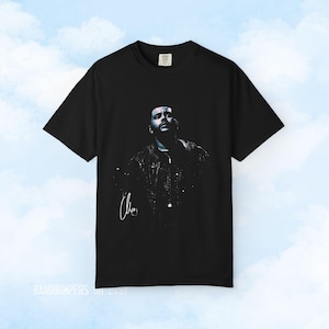 May include: A black short-sleeve t-shirt featuring a graphic print of a person with a dark, moody aesthetic. The design includes a signature in white script on the lower left side. The shirt has a standard crew neckline and a relaxed fit.