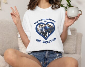 May include: White t-shirt with a blue heart-shaped graphic featuring a photo of a boy band and the text "my mom sold me to ONE DIRECTION". The shirt is paired with denim shorts.