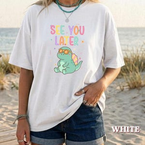 May include: A white short-sleeve t-shirt featuring a graphic of a light green dinosaur wearing orange sunglasses. Above the dinosaur, the text reads "SEE YOU LATER" in colourful, playful lettering. The design includes small decorative stars and squiggles.
