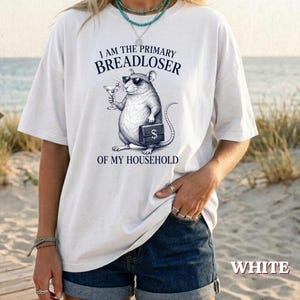 May include: A white short-sleeve t-shirt featuring a graphic of a rat wearing sunglasses, holding a martini glass and a briefcase. The text reads I AM THE PRIMARY BREADLOSER OF MY HOUSEHOLD. The shirt has a crew neckline and a loose, relaxed fit.