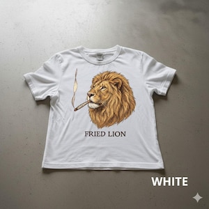 Può includere: T-shirt bianca con un'illustrazione dettagliata di un leone che fuma un sigaro. Il leone ha una criniera marrone e sta fumando un sigaro acceso. Il testo "FRIED LION" è stampato sotto la grafica del leone. La parola "WHITE" è stampata in basso.