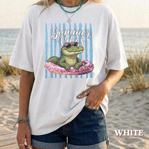 Può includere: Una t-shirt bianca con la grafica di un alligatore verde che indossa occhiali da sole mentre galleggia su un salvagente rosa. Il design include strisce verticali blu e la scritta Summer Vibes in corsivo. La maglietta ha maniche corte.